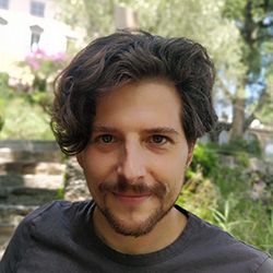Dragan Ahmetovic, a white man with brown hair and short beard, wearing a gray t-shirt, looking at the camera and smiling, with a garden and a building out of focus in the background.