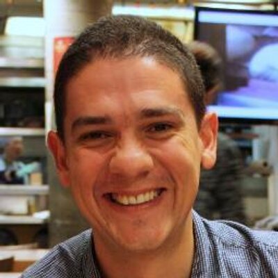 The image of John Guerra Gomez, a brown skinned man with short hair smiling at the camera