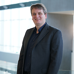 Christian Vogler, a white man with dark blonde short hair, is wearing a dark shirt and a dark jacket, standing against a brightly lit row of windows