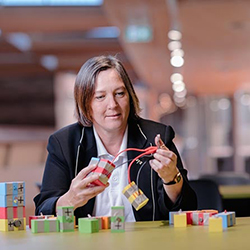 Kirsten Ellis, White women with bobbed brown hair sitting at a desk looking at the foam blocks and wires she is holding