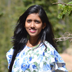 Taslima Akter is smiling at the camera. She is a brown skinned woman with black long straight hair. She is wearing white and blue floral-printed dress. Background is blurry and contains green trees. 