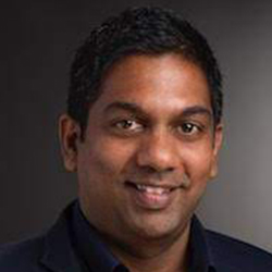 Roshan Peiris, a south asian man with black hair wearing a dark blue suit with a dark blue open shirt in front of a dark background