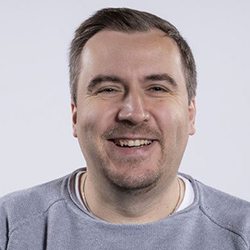 Benjamin Tannert, a white man with short brown hair, wearing a grey sweater and smiles at the camera infront of a white background