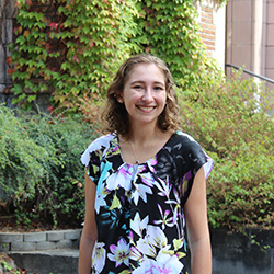 Kelly Mack, a white woman in her 20s with curly blondish brown hair and a floral blouse stands in front of autumn changing leaves on UW campus