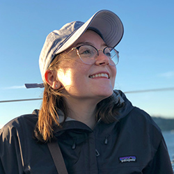 Jaylin Herskovitz is a white woman with brown hair, sitting on a boat, she wears a blue jacket and blue baseball cap