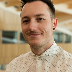 Kyle Montague, a white man with short brown hair wearing a white shirt with black polka dots, smiling at the camera with an out of focus building interior in the background