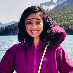 Image of Sowmya Somanath, a south asian woman with black hair, wearing a purple jacket, standing on a ferry with a background of mountains and ocean, and smiling at the camera